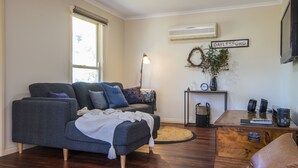 A living room with a grey sofa, a small wooden table, a wall-mounted air conditioner, and a sign that reads "DAYLESFORD". - Birdie (Daylesford)
