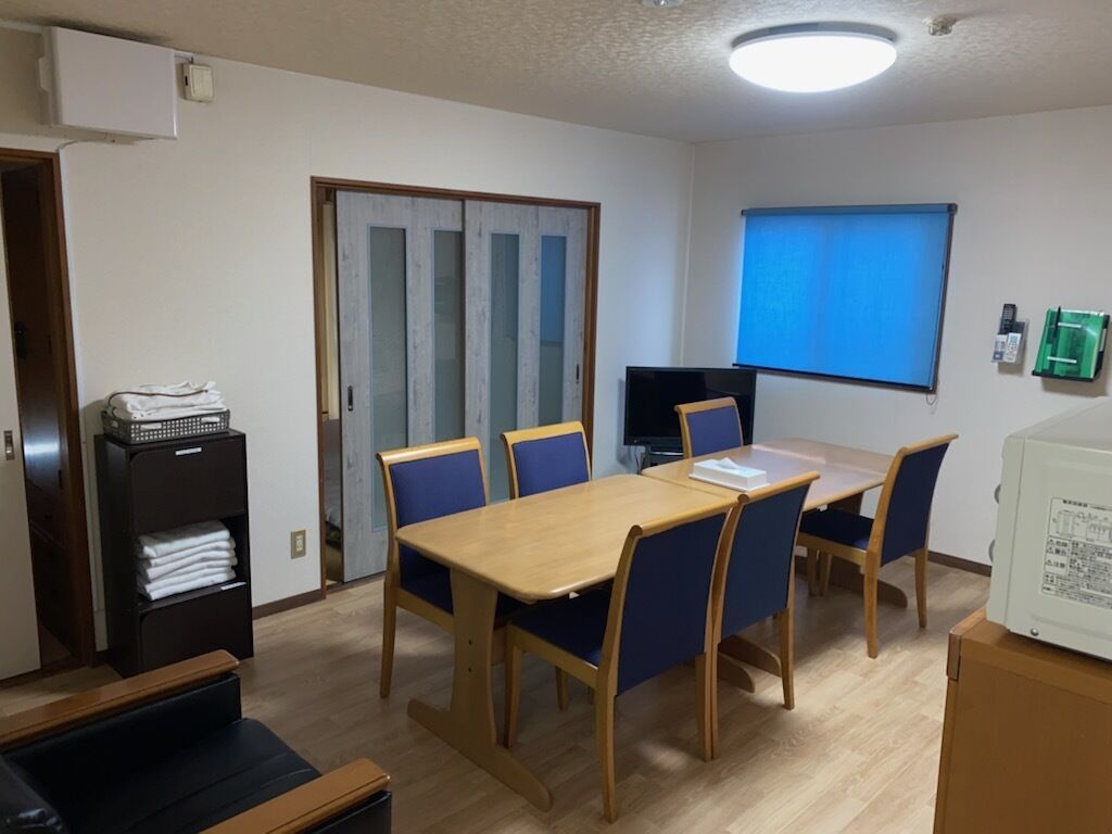 Ein Zimmer mit Holztisch, blauen Stühlen, einem Fernseher, einem Fenster mit Jalousien, einer Tür und einer kleinen Küchenzeile.