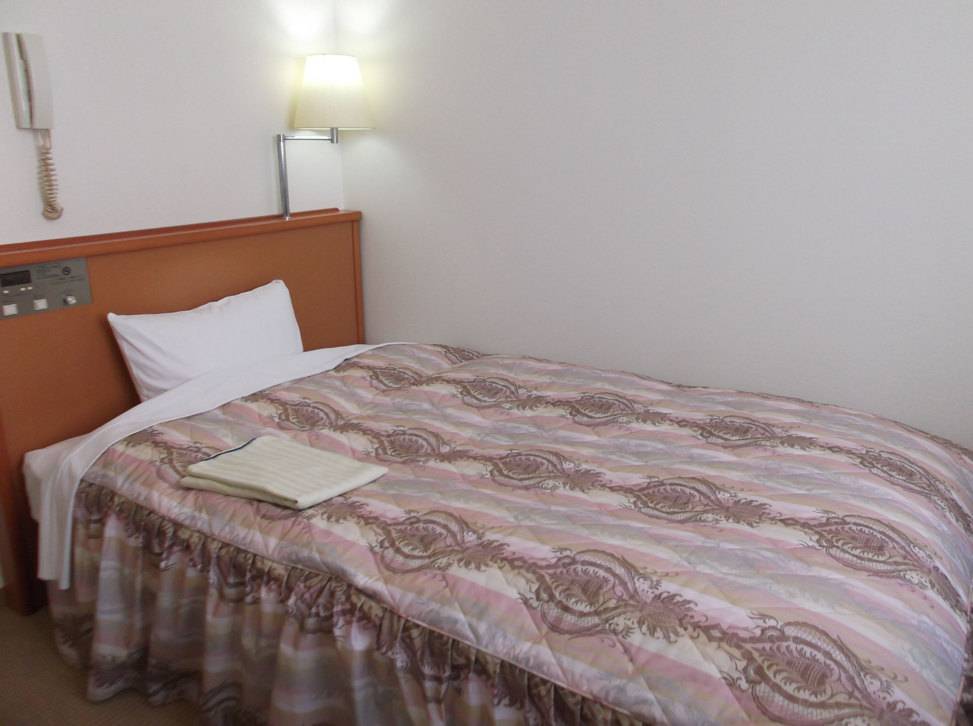 A neatly made bed with a patterned bedspread and a white pillow, a bedside lamp, and a control panel on the wall.