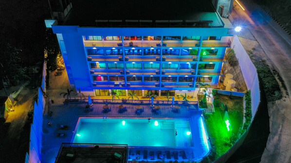 Ein mehrstöckiges Hotel mit Poolbereich auf dem Dach, Außenbereich zum Sitzen und einer gut beleuchteten Fassade.