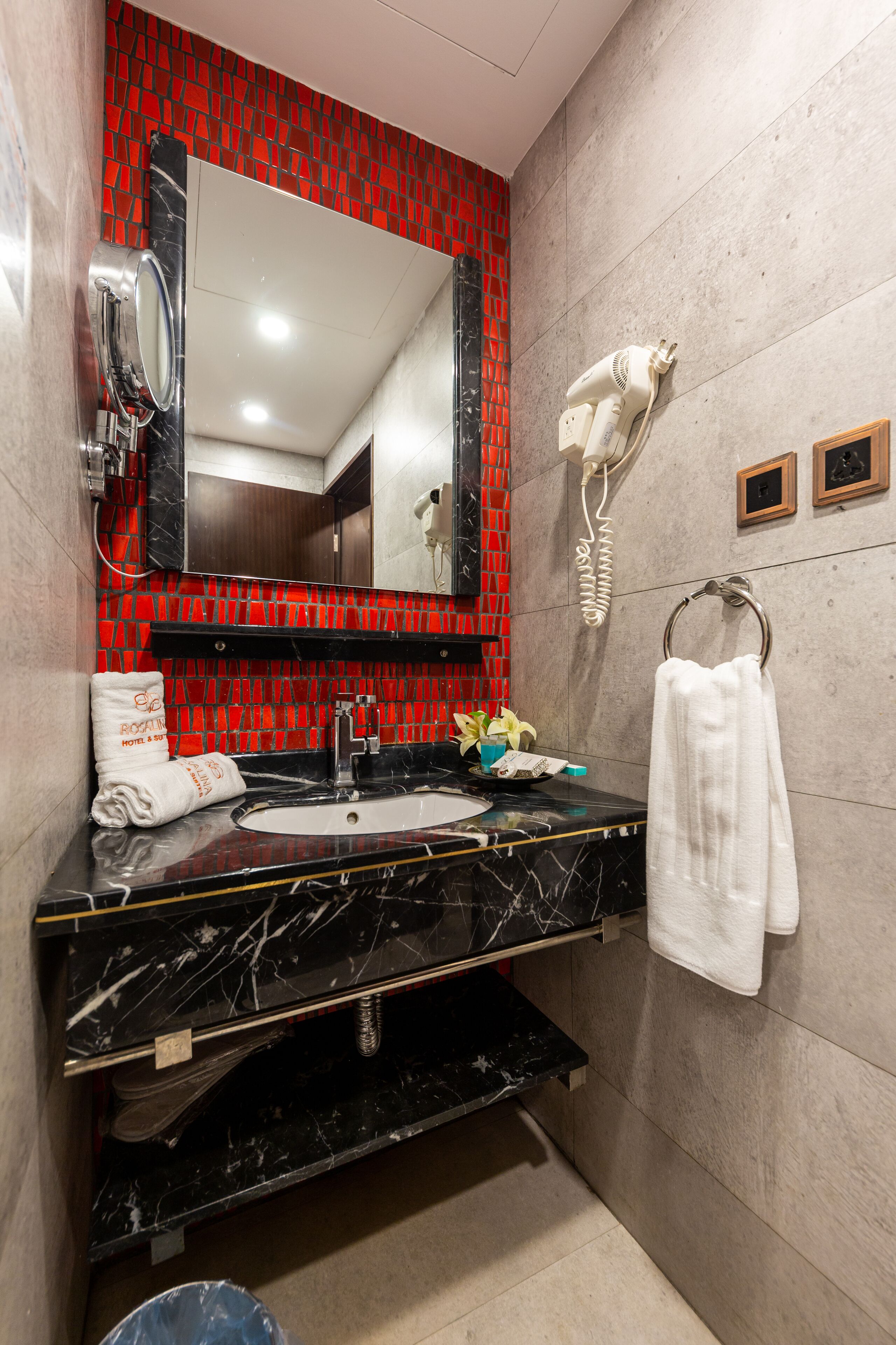 A bathroom with a black marble sink, a red mosaic wall, a white towel, and a hairdryer on the wall.