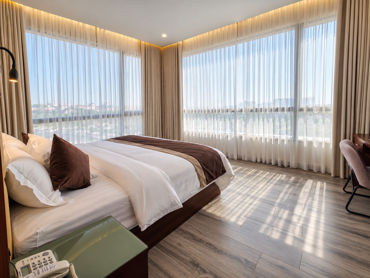 A hotel room with a large bed, a desk, and a view of a cityscape through sheer curtains.