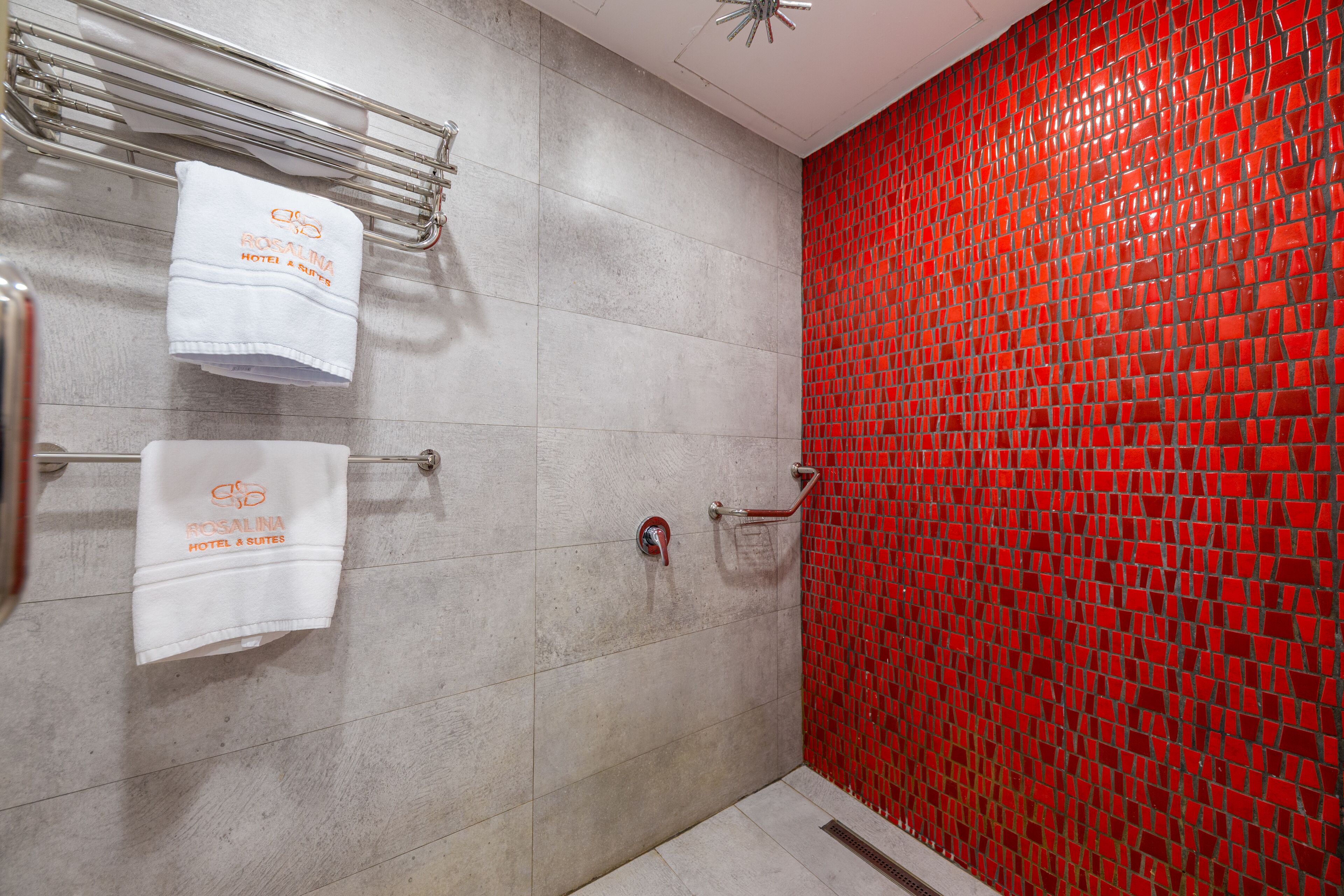 Un baño con pared de mosaicos rojos, una ducha con puerta de cristal y dos toallas colgadas en un tendedero.