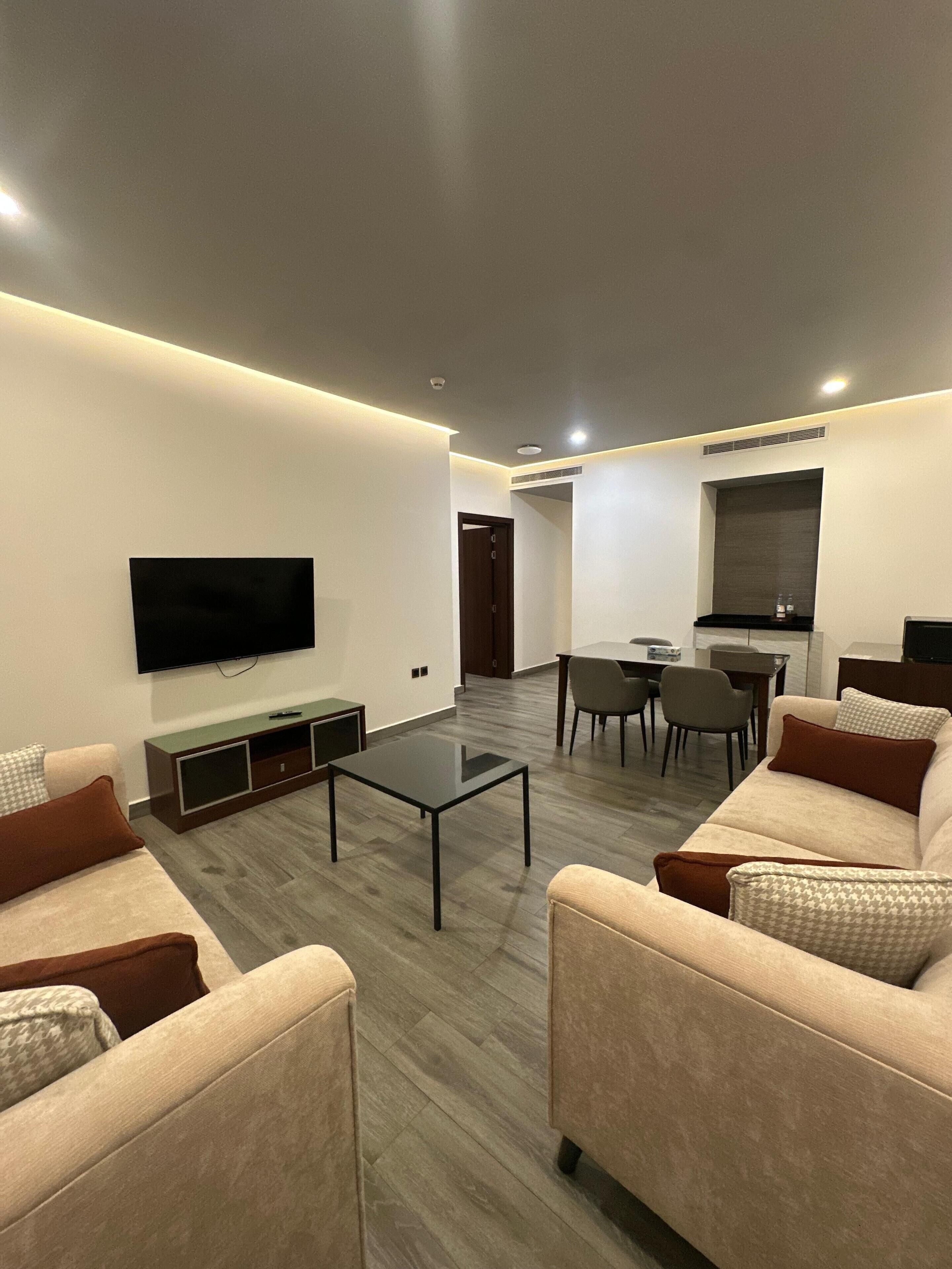 A modern living room with a flat-screen TV, a coffee table, sofas, and a dining area with chairs.