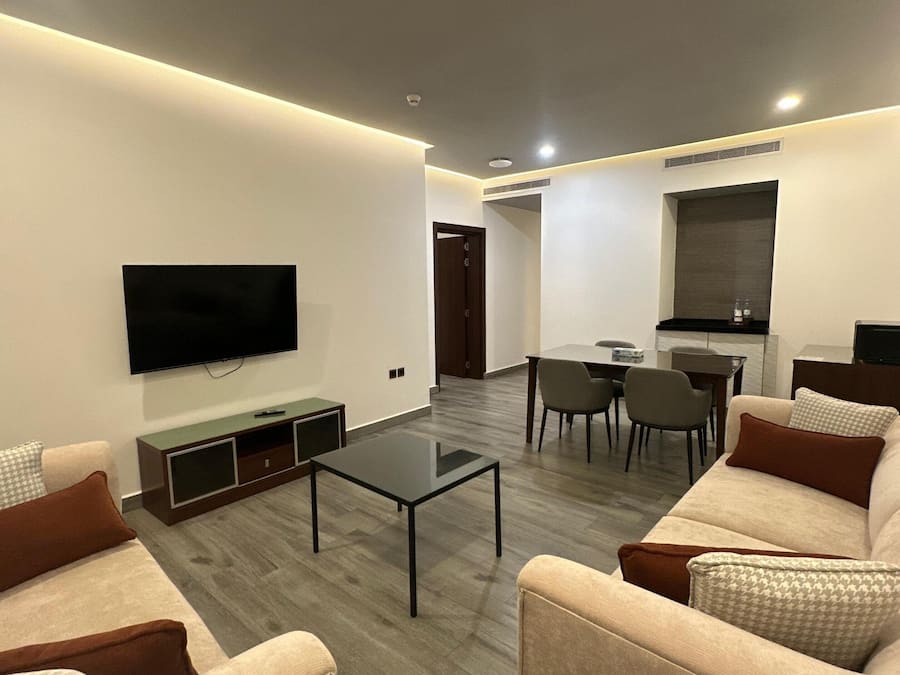 A modern living room with a flat-screen TV, a coffee table, sofas, and a dining area with chairs.