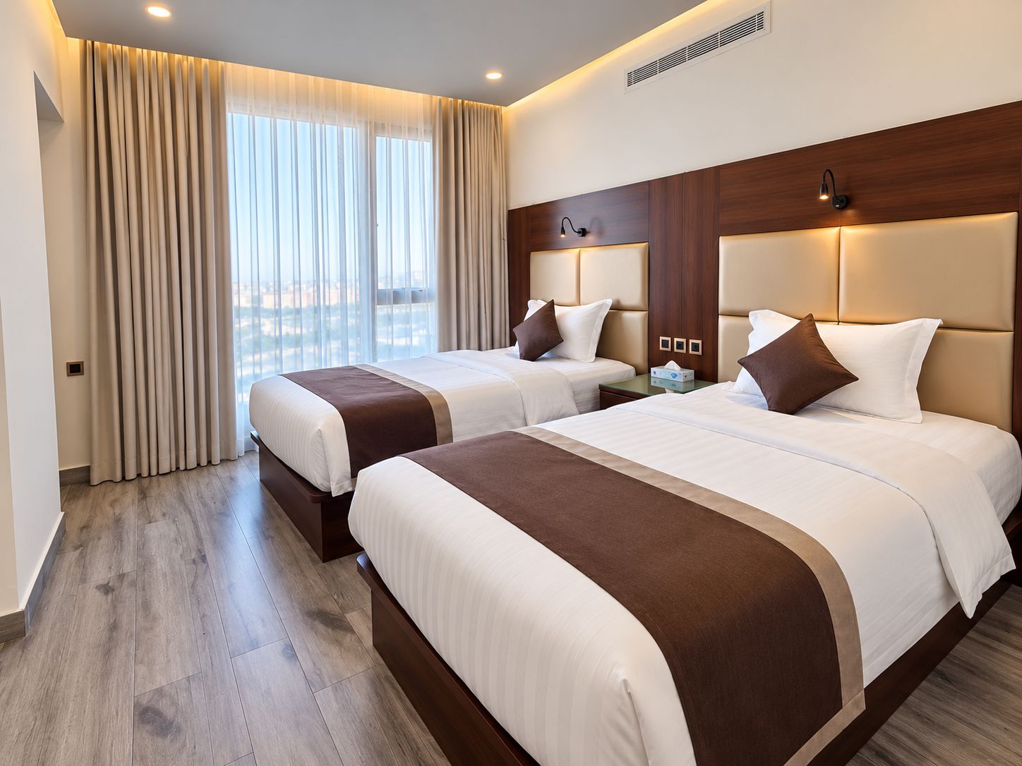 A hotel room with two beds, a large window with curtains, a wooden headboard, and a nightstand with a lamp.