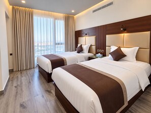 A hotel room with two beds, a large window with curtains, a wooden headboard, and a nightstand with a lamp.