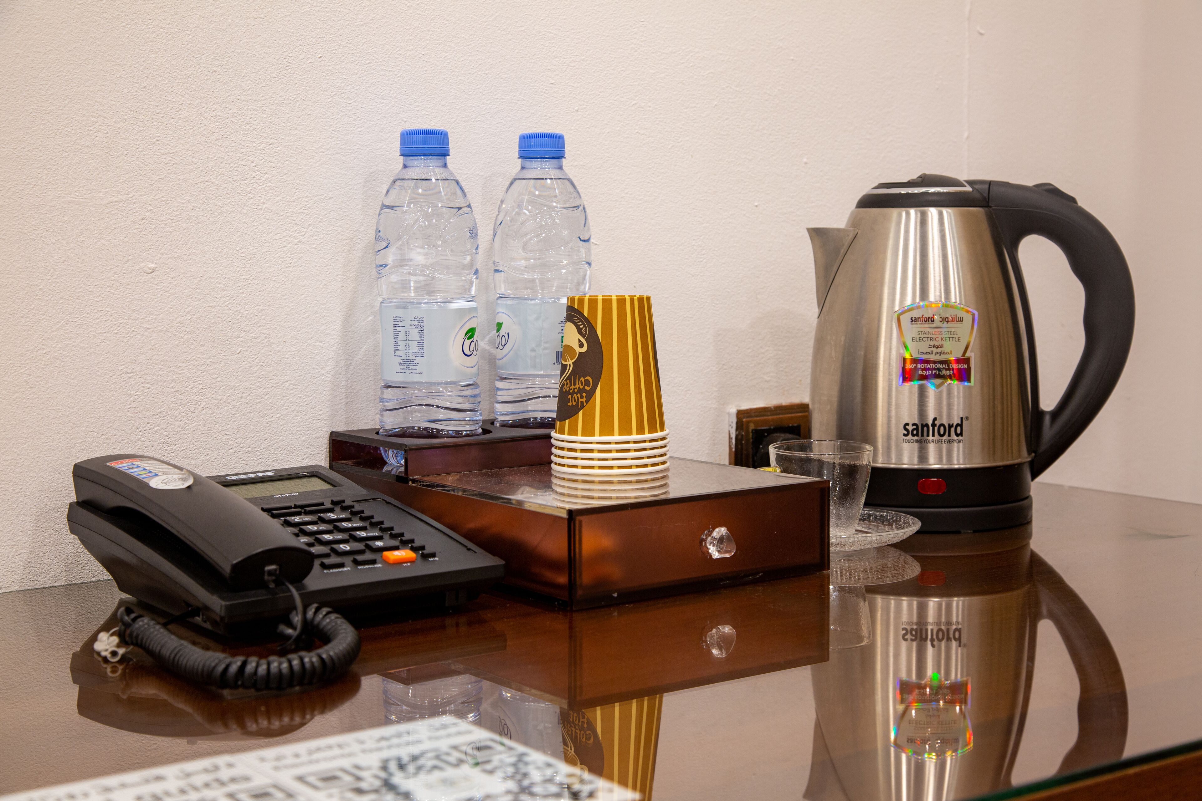 Un mostrador de recepción de hotel con un teléfono, dos botellas de agua, una pila de platos amarillos y blancos, un hervidor eléctrico de acero inoxidable y un vaso.