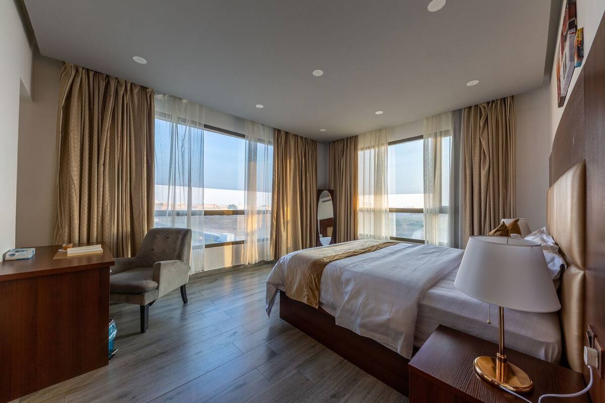 A hotel room with a bed, desk, chair, and large windows with curtains.