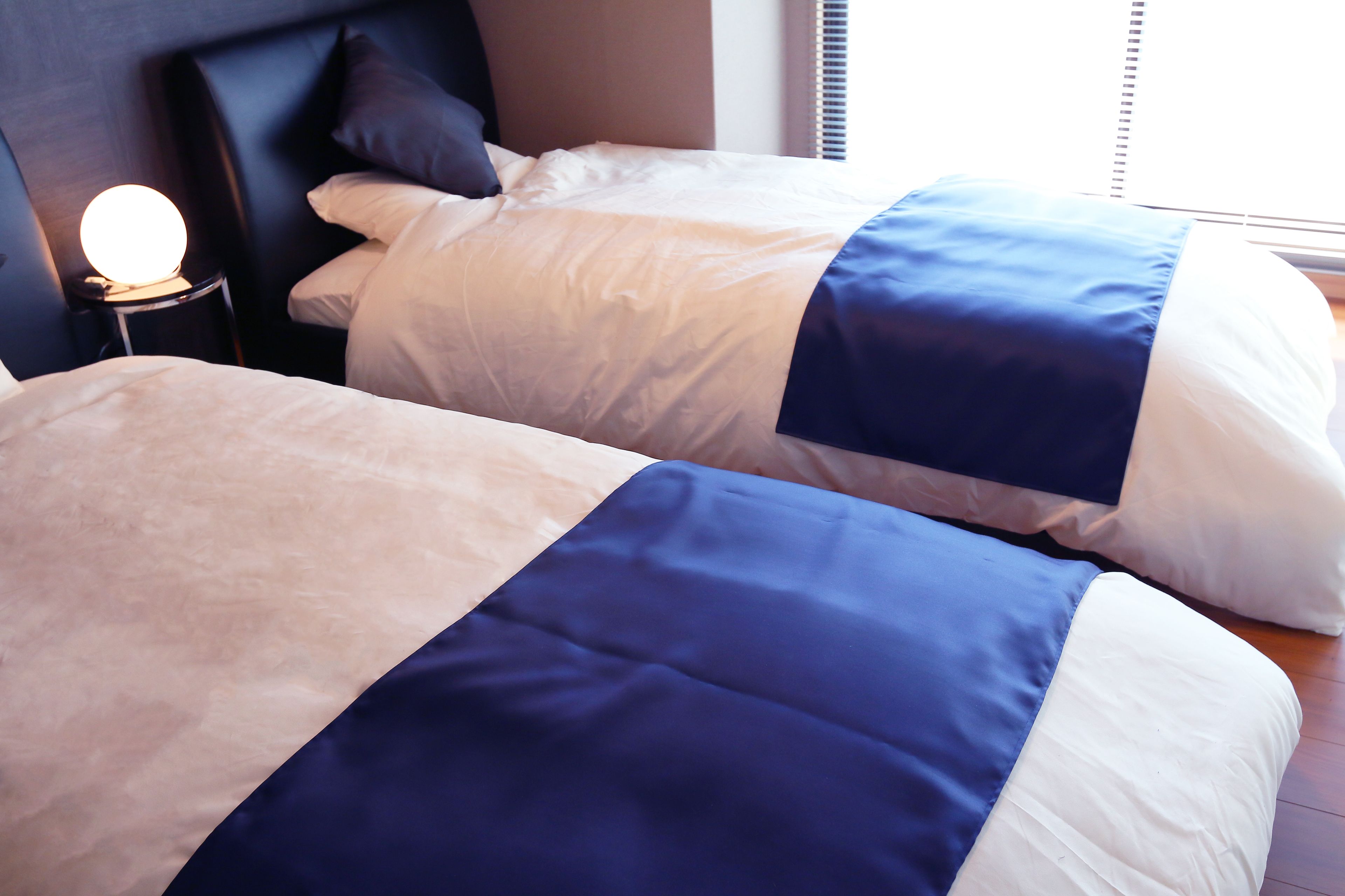 A hotel room with two beds, a bedside table with a lamp, and a window with blinds.