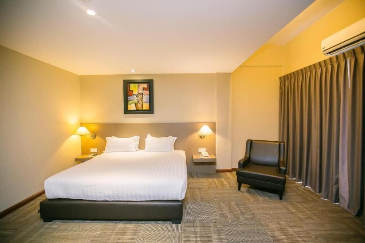 A hotel room with a large bed, two bedside lamps, a chair, a painting on the wall, and an air conditioning unit.