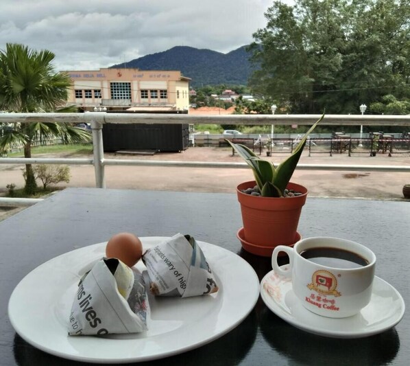 A table with a plate of food, a cup of coffee, and a potted plant on a balcony overlooking a building and mountains.
