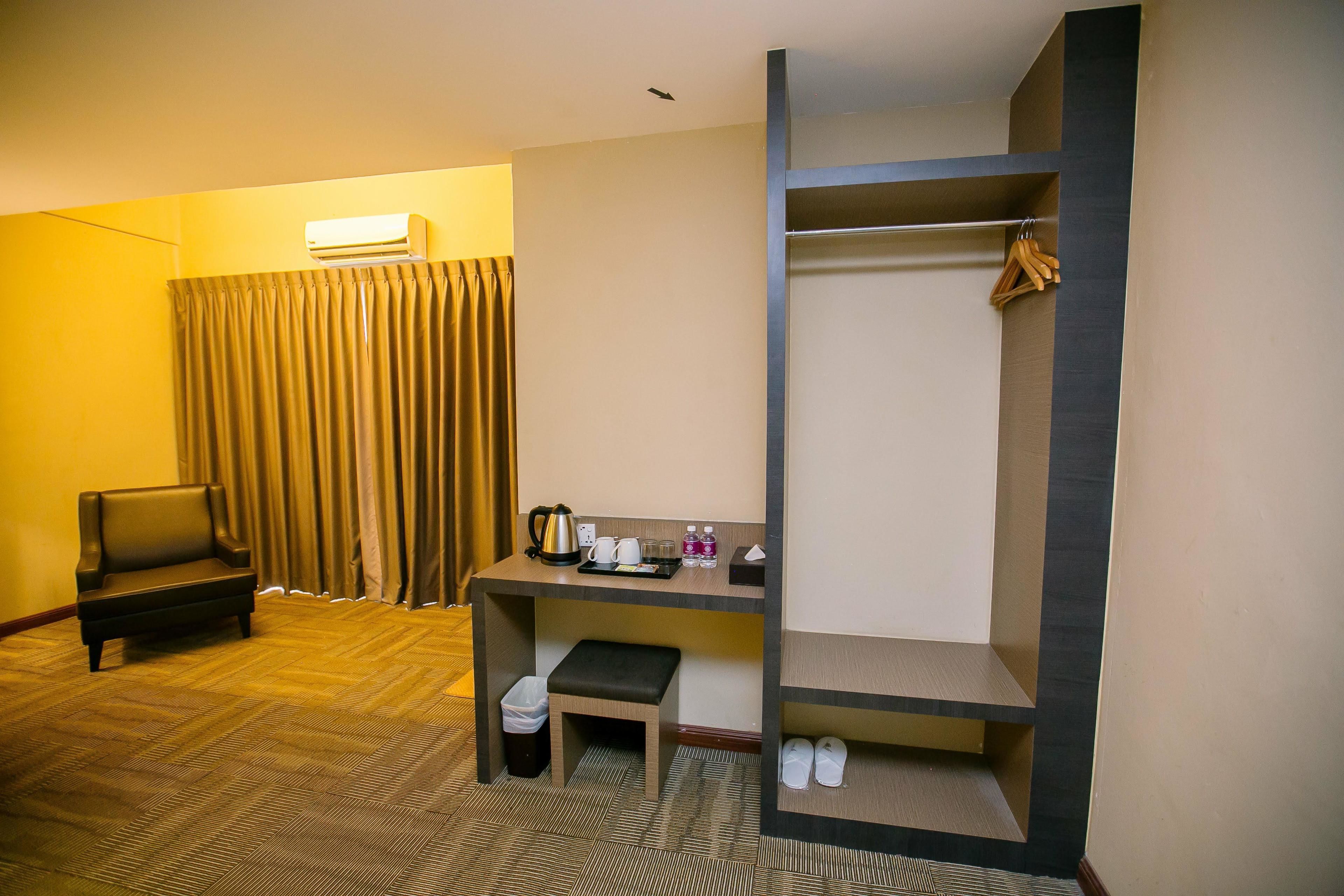 A hotel room with a sofa, a desk with a kettle, a bench, a coat rack, and a trash bin.
