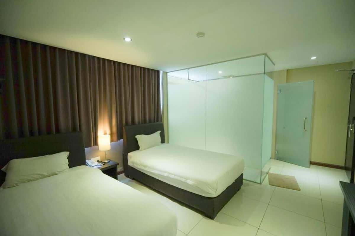 A hotel room with two beds, a nightstand with a lamp, a wardrobe, and a shower area.