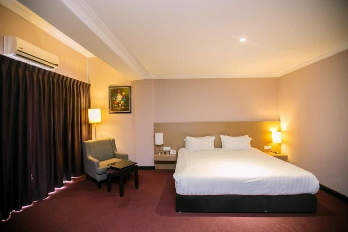 A hotel room with a large bed, a desk, a chair, an air conditioning unit, and a nightstand with a lamp.