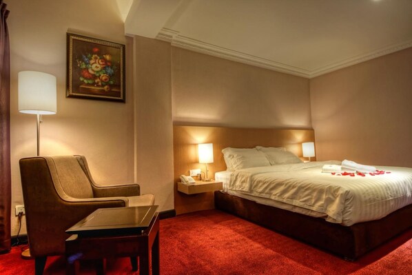 A hotel room with a bed, a chair, a table, a nightstand with a lamp, and a painting on the wall.