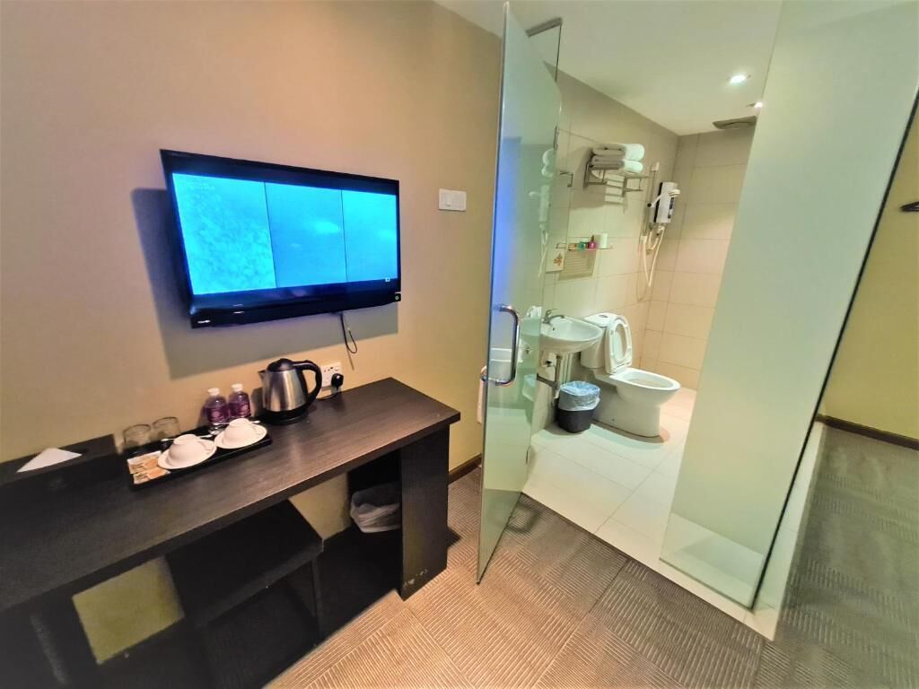 A modern bathroom with a glass shower enclosure, a sink, and a toilet. There is a flat-screen TV mounted on the wall and a small table with a kettle and cups.