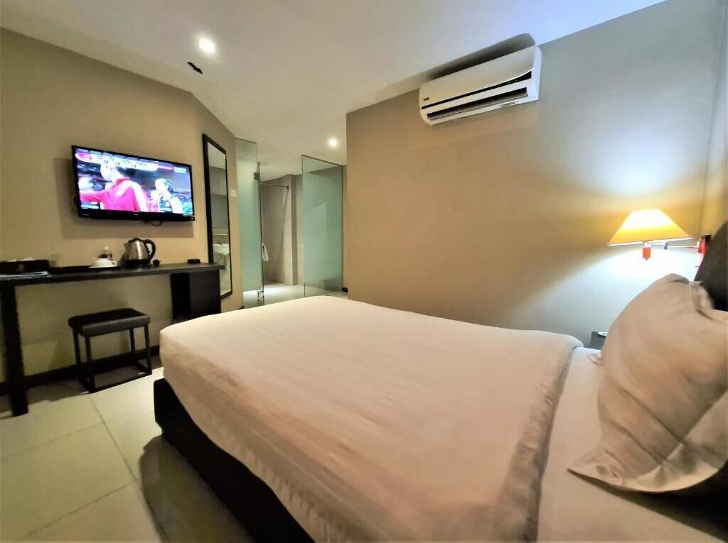 A hotel room with a bed, a TV mounted on the wall, a desk with a chair, and an air conditioning unit.