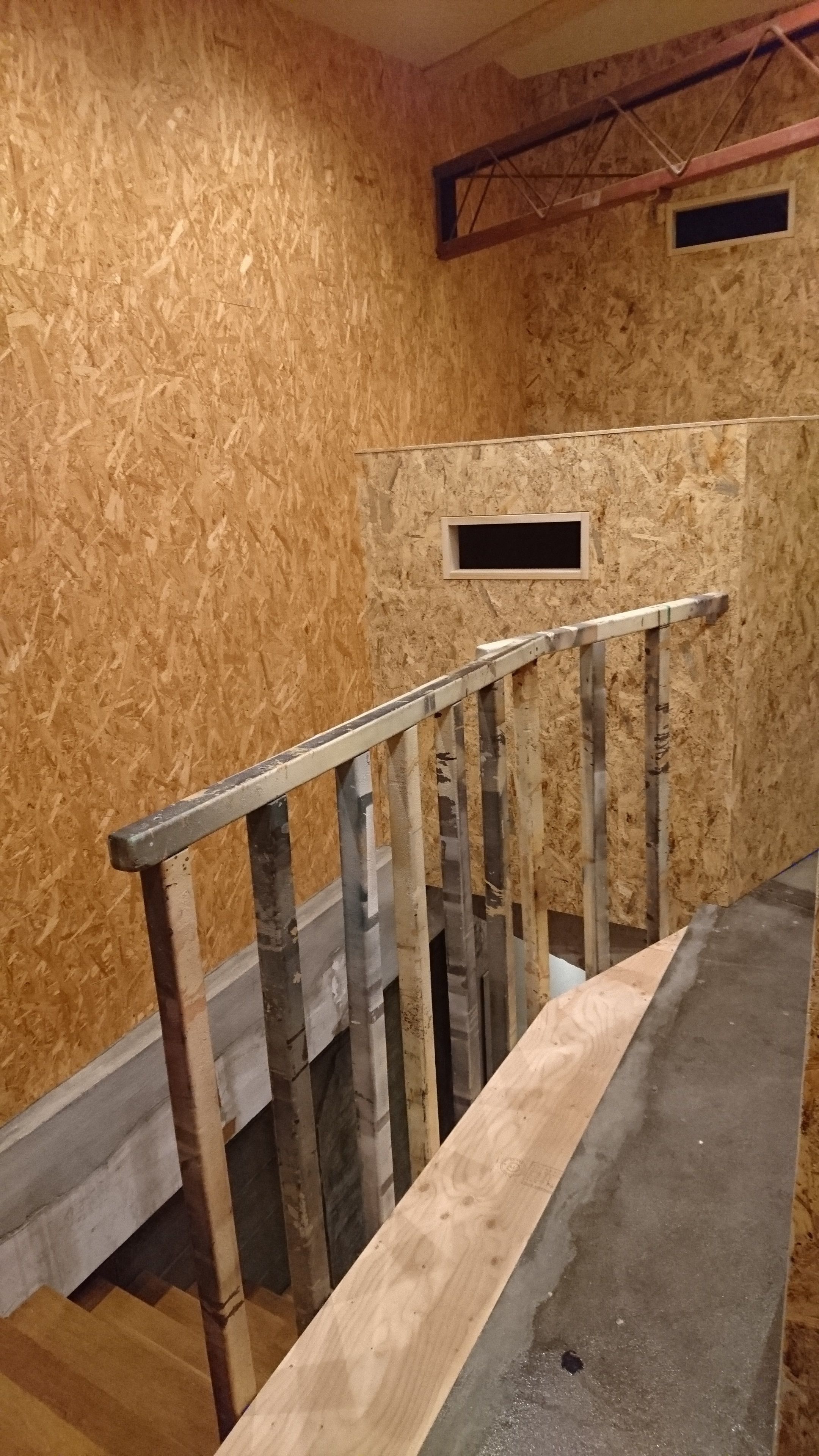 Una escalera de madera con barandales de metal, un rellano de hormigón y una pared parcialmente terminada con montantes de madera a la vista.