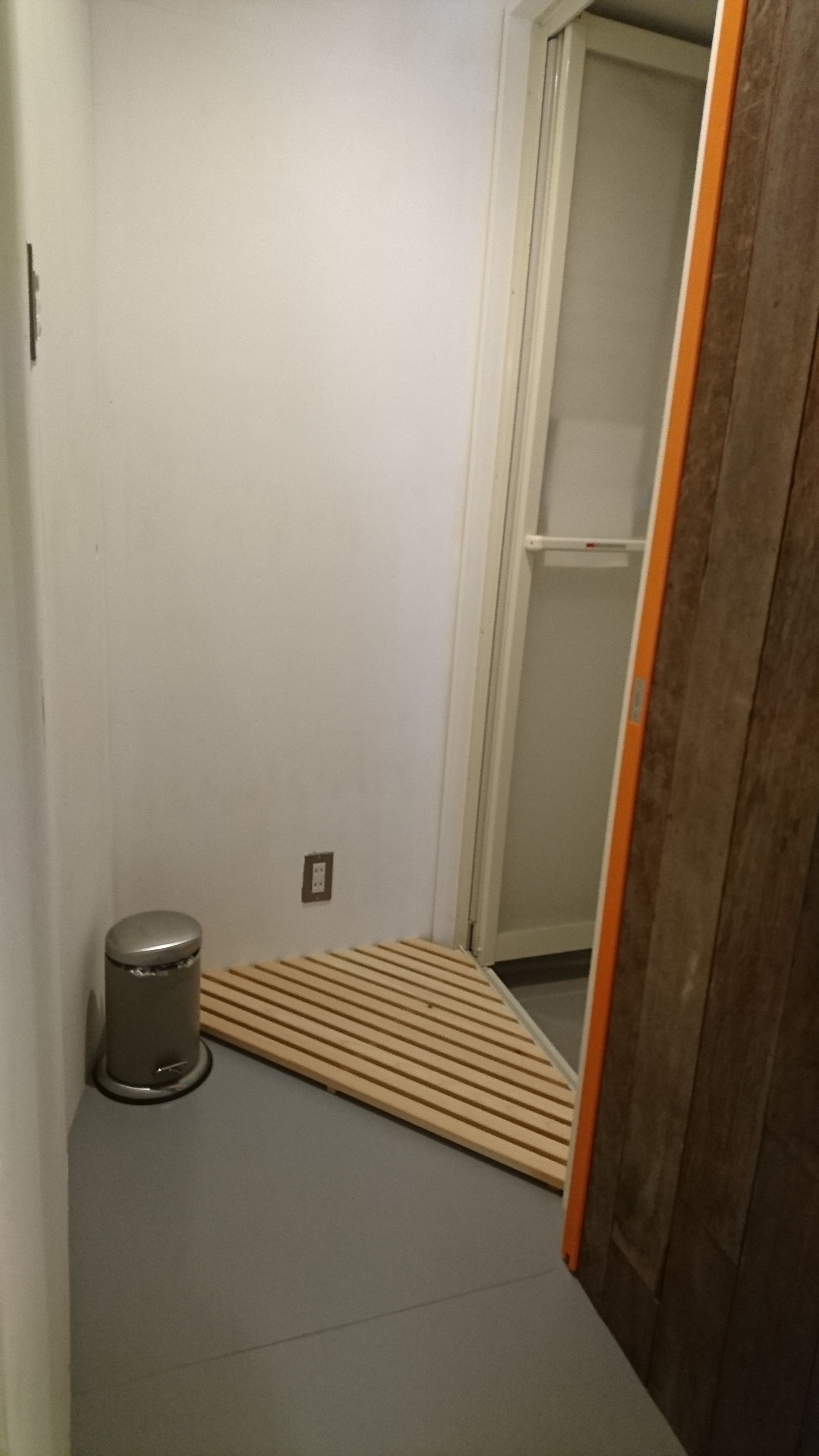 Una habitación pequeña y estrecha con piso gris, un banco de madera con listones y un cubo de basura pequeño, además de una puerta que da a otra habitación.