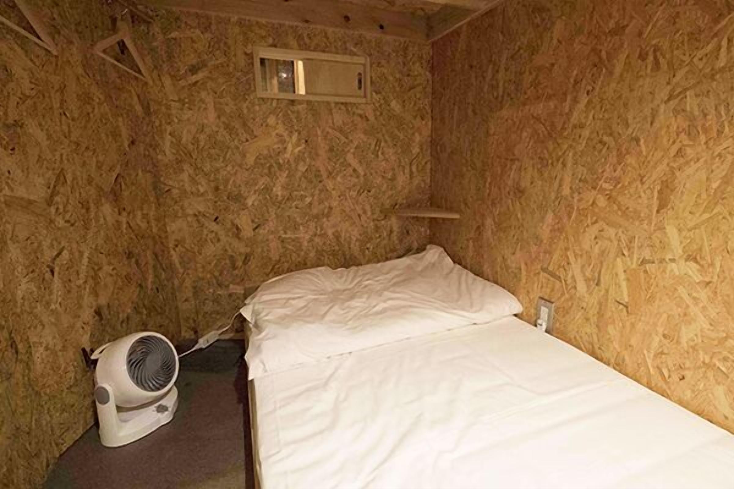 A small, sparsely furnished room with a single bed, a white fan, and a small shelf.