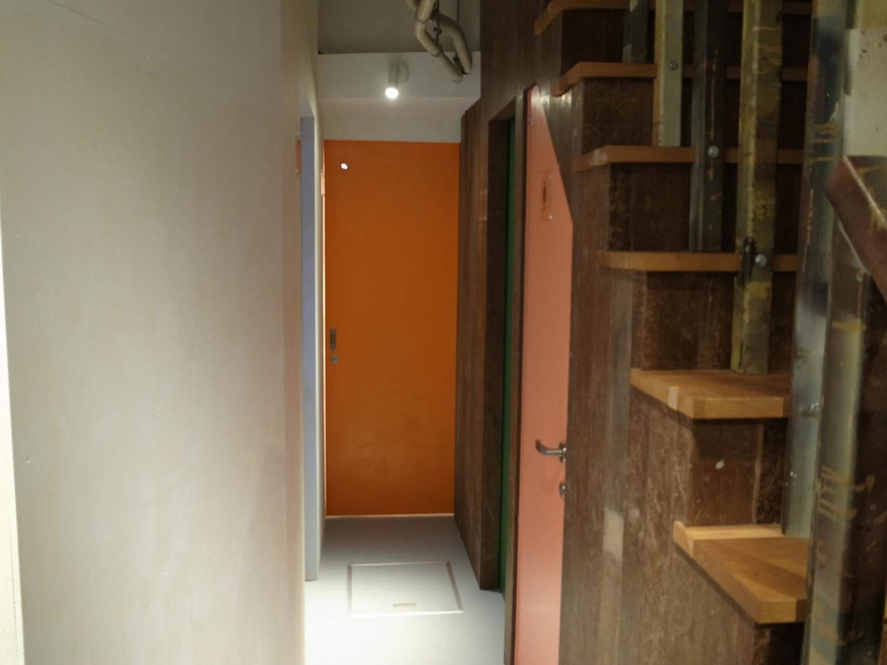 A narrow hallway with a wooden staircase on the right and an orange door at the end.