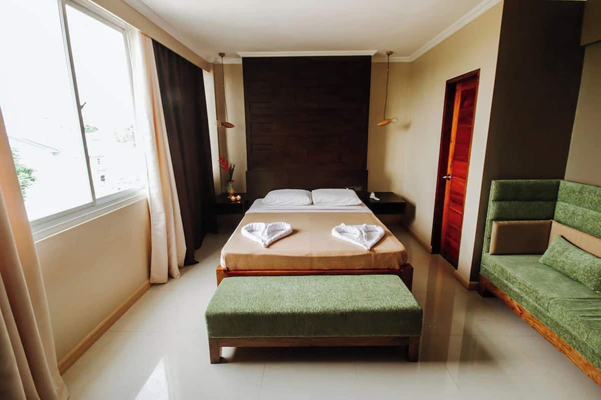 Habitación de hotel con una cama, un banco verde, una mesita de noche, una lámpara y vistas al exterior.
