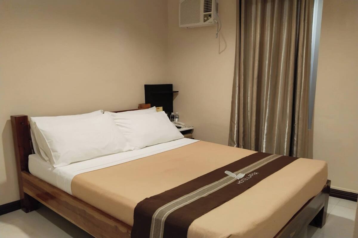 Una cama bien hecha con sábanas blancas y colcha marrón, una mesita de noche con lámpara, y una ventana con cortinas.