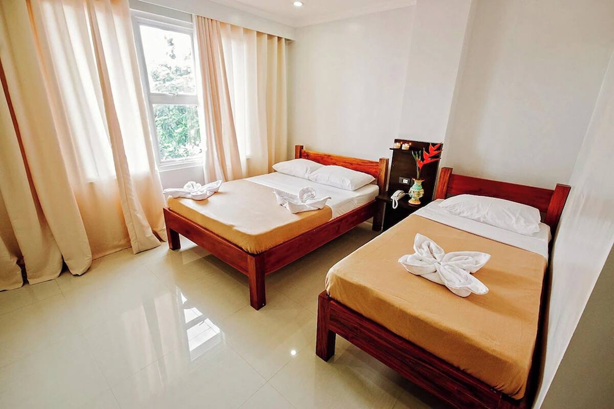 Habitación de hotel con dos camas de madera, cortinas color beige, una ventana con vistas a la vegetación y una mesita de noche con un florero.