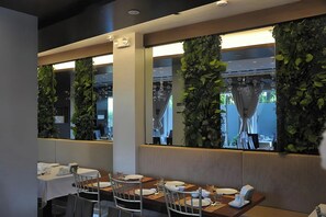 A restaurant with a long table, chairs, and a view through a window with greenery.