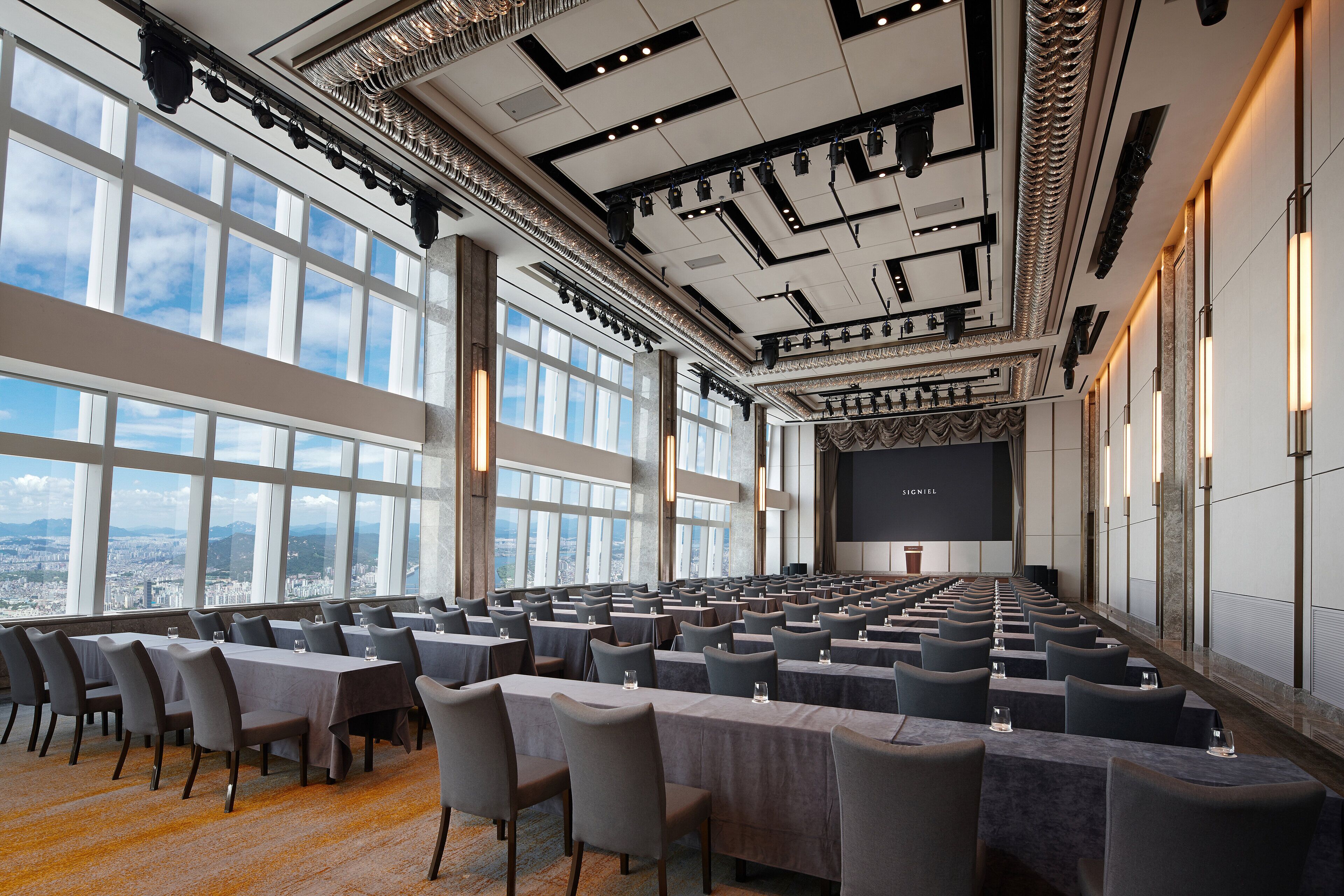A conference room with rows of tables and chairs, large windows with a city view, and a stage with a screen.