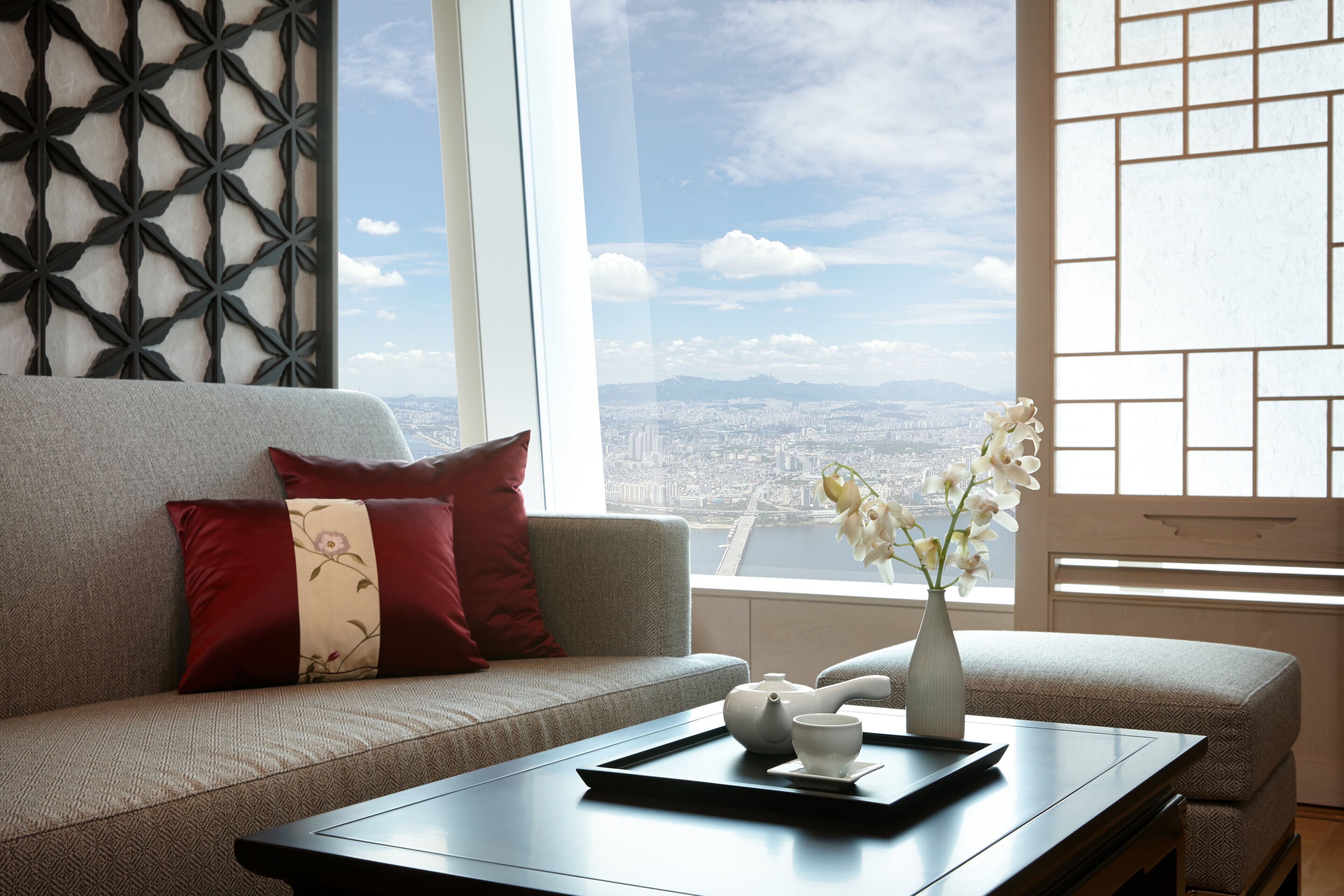 A modern living room with a sofa, a coffee table, a vase with flowers, and a view of a cityscape through large windows.