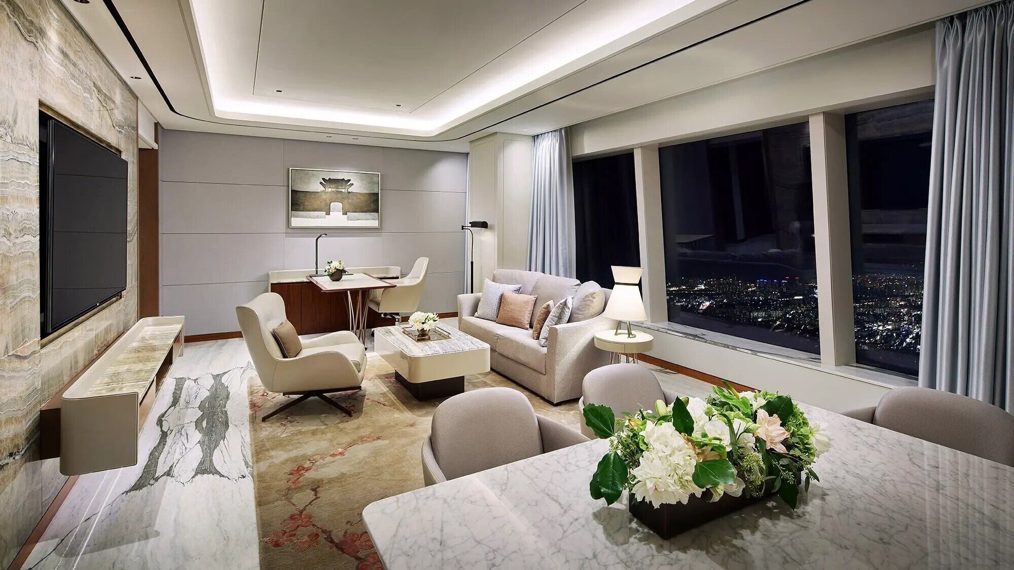 Una sala de estar moderna con un sofá, sillones, una mesa de centro y una mesa de comedor con un centro de mesa floral. Hay un cuadro en la pared, un televisor montado y un ventanal con vistas a la ciudad.