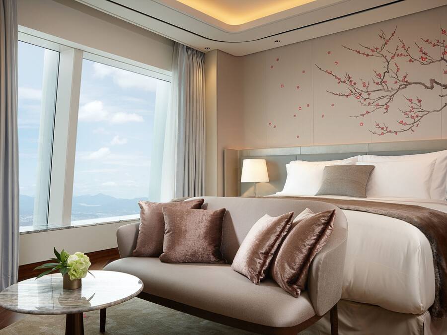 A modern hotel room with a large bed, a sofa, a round table with a vase, and a view of the city through large windows.