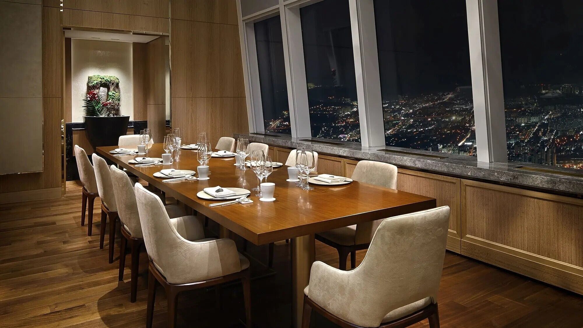 A dining area with a long table set for a meal, large windows with a city view, and a decorative plant in the corner.