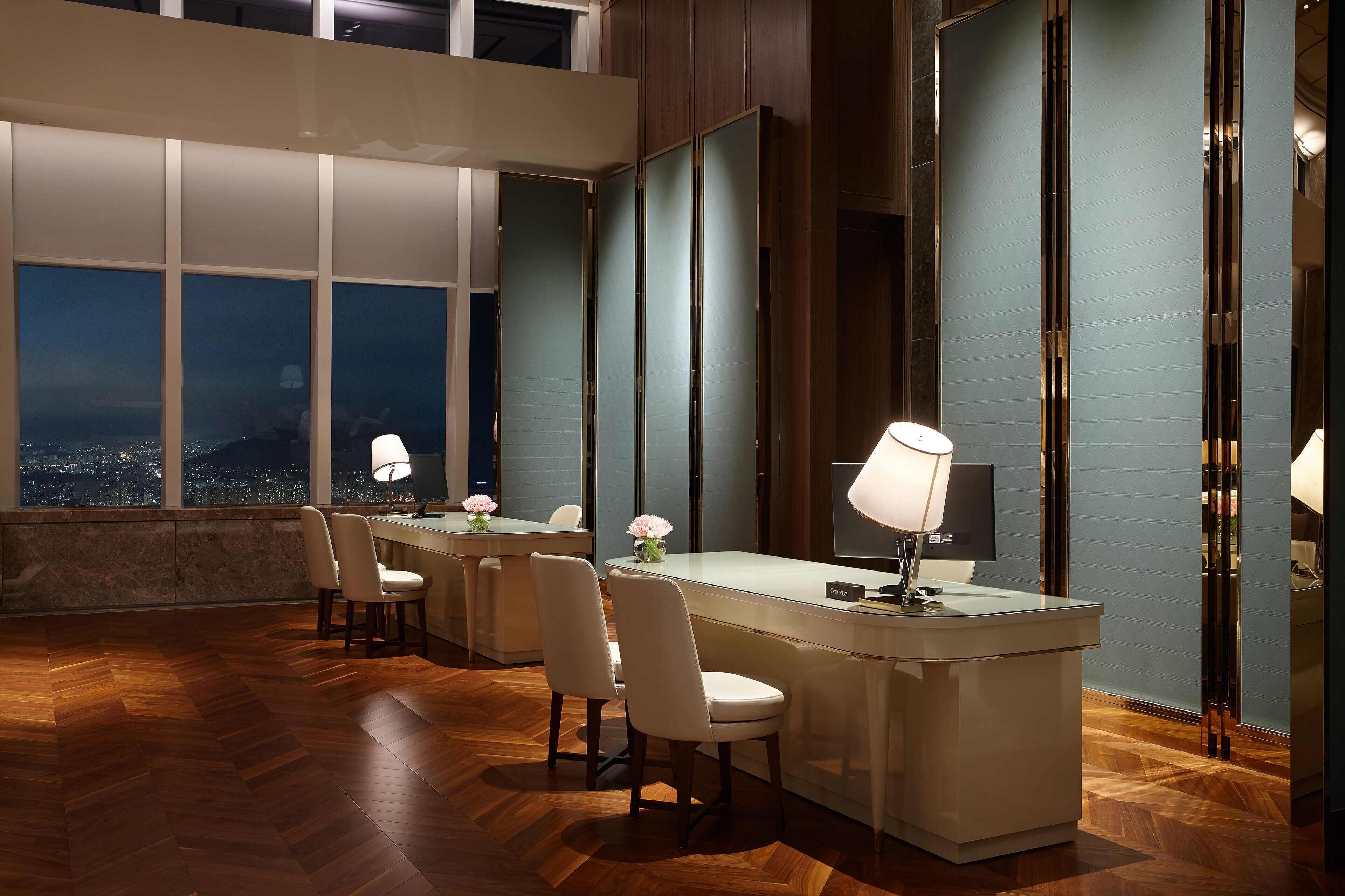 A modern hotel room with a large window offering a city view, a wooden herringbone floor, a central desk with chairs, and a decorative vase with flowers.