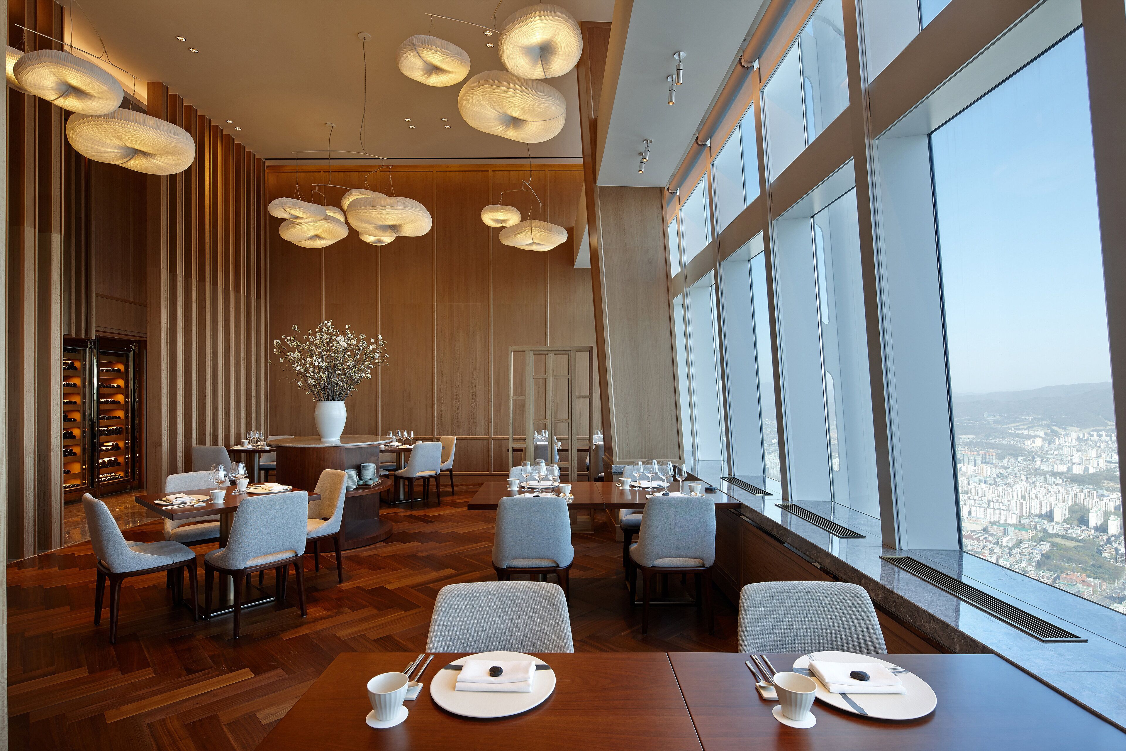 A modern dining area with high ceilings, large windows, and a city view.