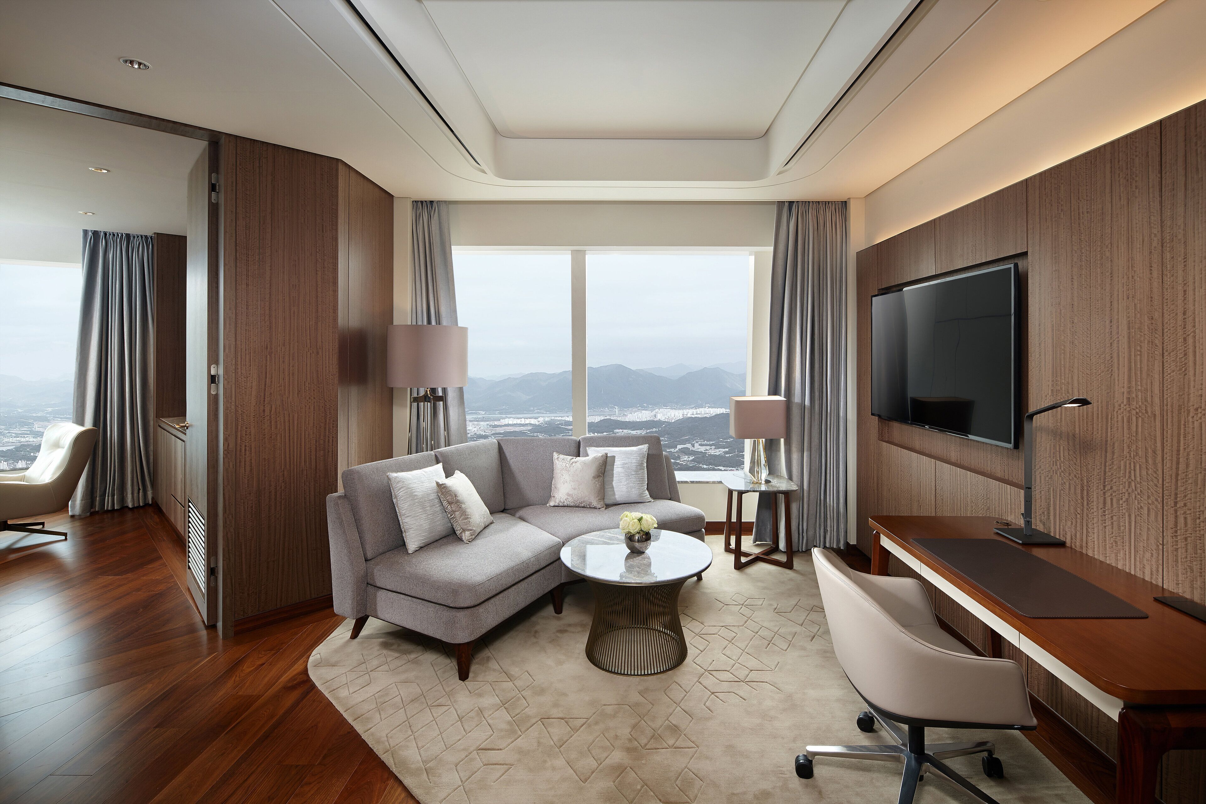 A modern hotel room with a large window offering a city view, a gray sectional sofa, a round coffee table, a desk with a chair, and a flat-screen TV mounted on the wall.