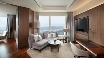 A modern hotel room with a large window offering a city view, a gray sectional sofa, a round coffee table, a desk with a chair, and a flat-screen TV mounted on the wall.