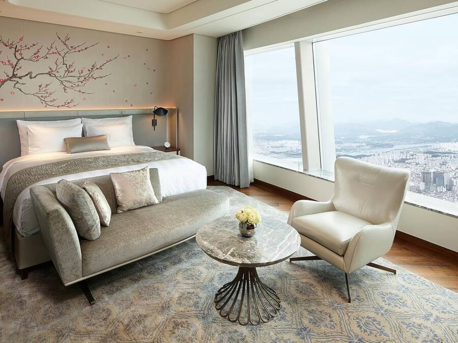 Ein modernes Hotelzimmer mit einem großen Bett, einer Couch, einem Sessel und Blick auf eine Stadtlandschaft durch große Fenster.