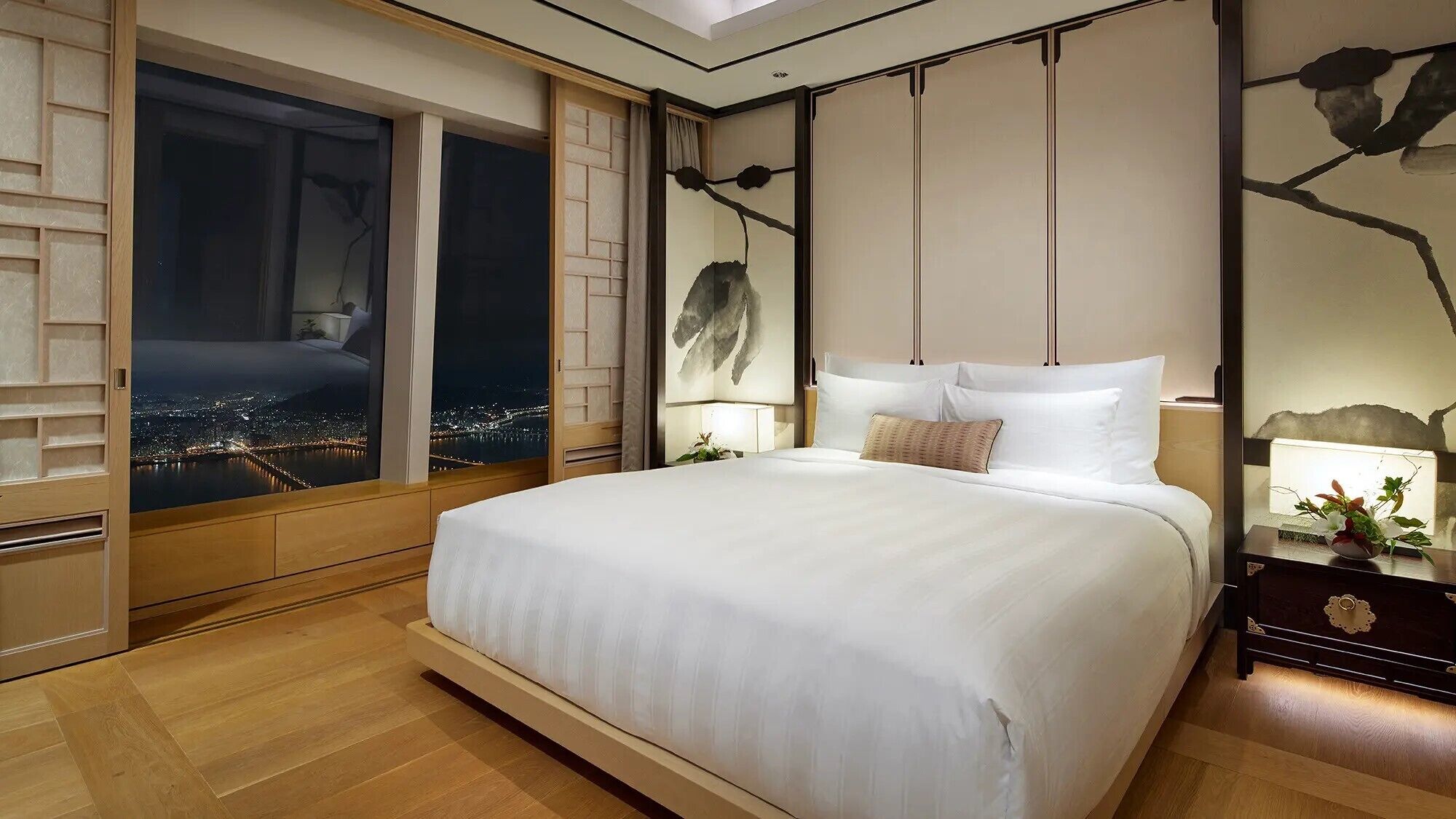Une chambre moderne avec un grand lit, une vue nocturne sur le paysage urbain et un mur décoratif orné de motifs représentant des branches.