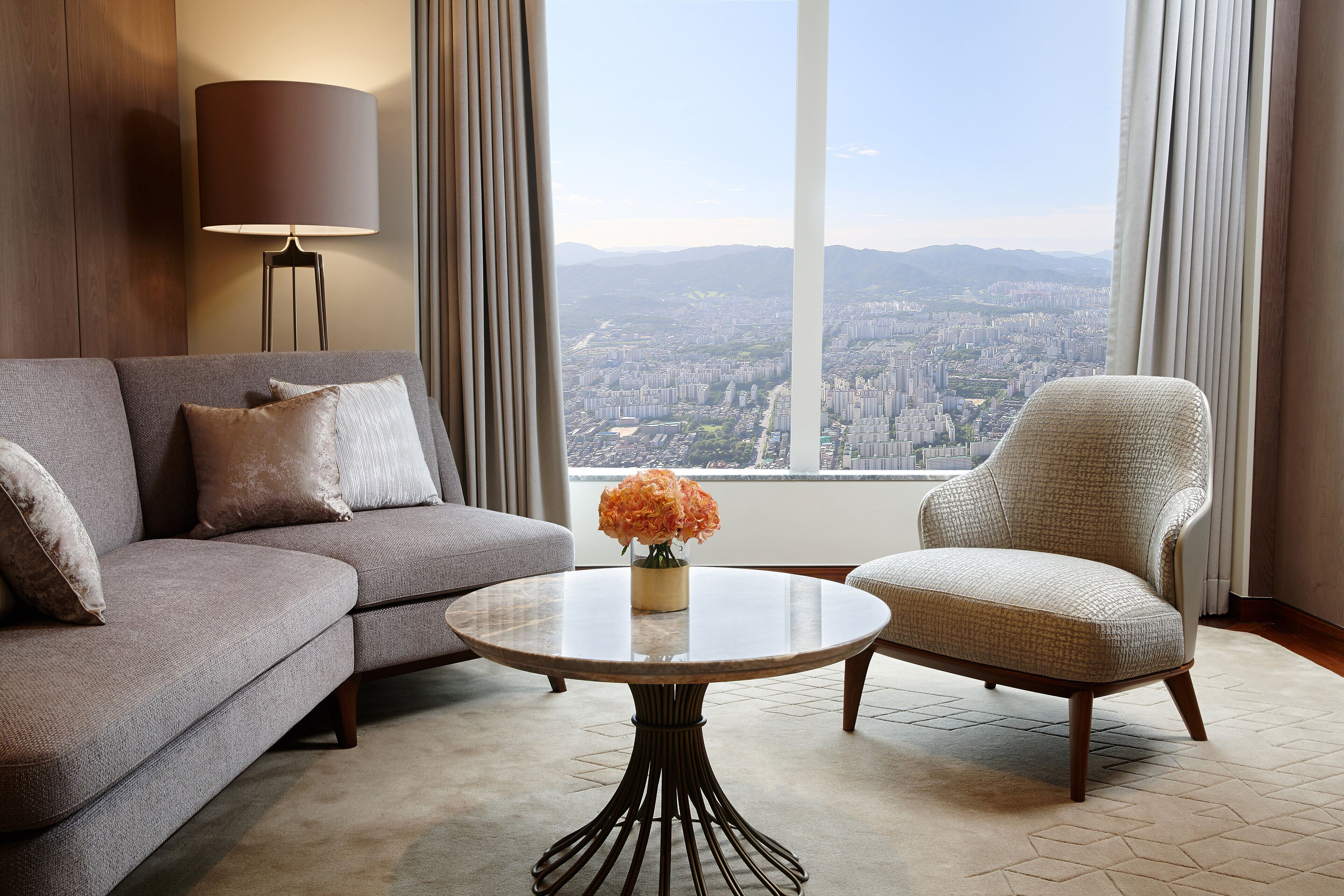 A modern living room with a sofa, armchair, and round coffee table. A lamp is on the left, and a view of a cityscape is visible through the window.