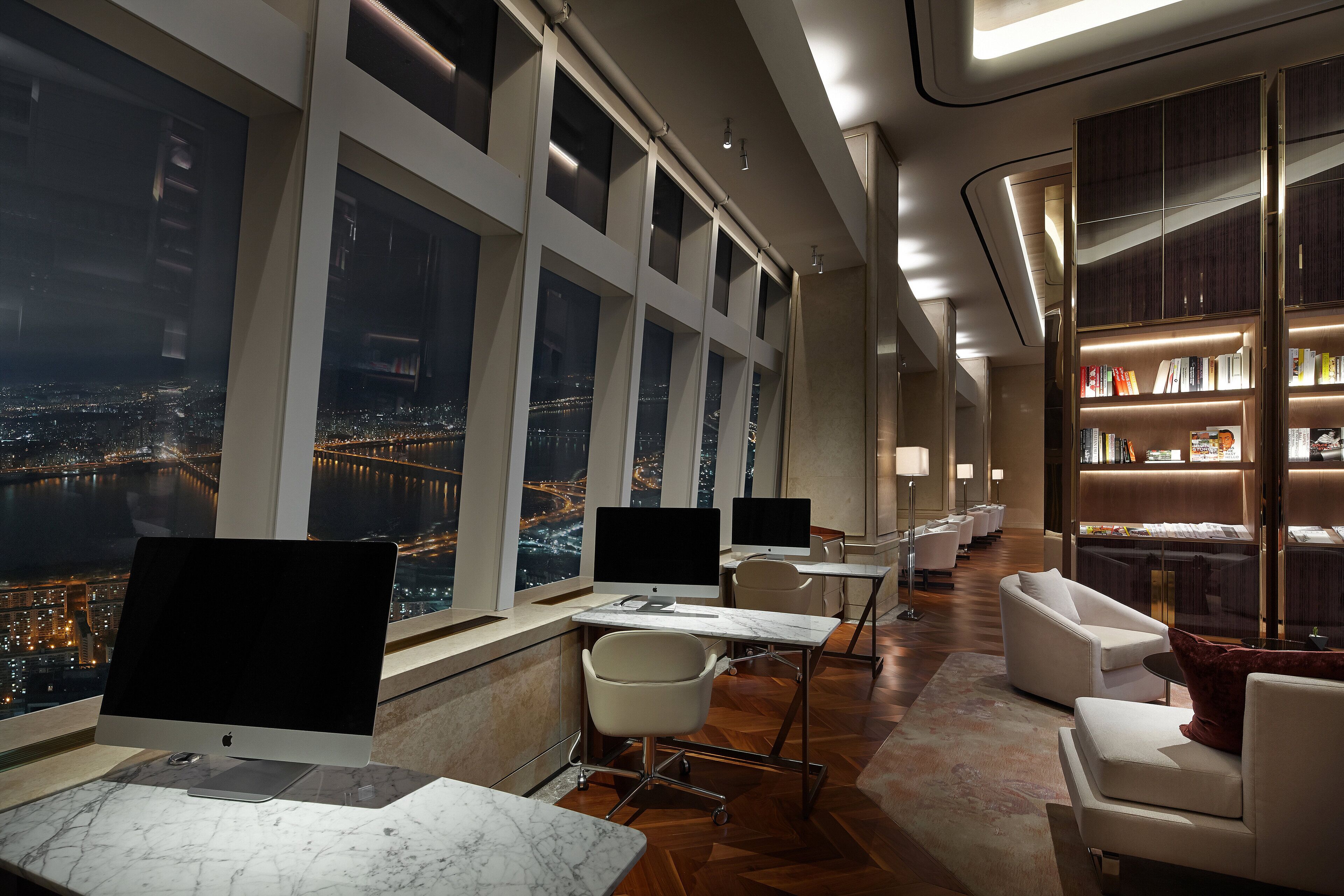 A modern hotel room with large windows, a desk with a computer, a comfortable seating area, and a bookshelf.