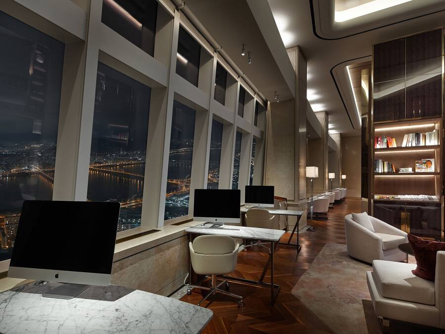 A modern hotel room with large windows, a desk with a computer, a comfortable seating area, and a bookshelf.