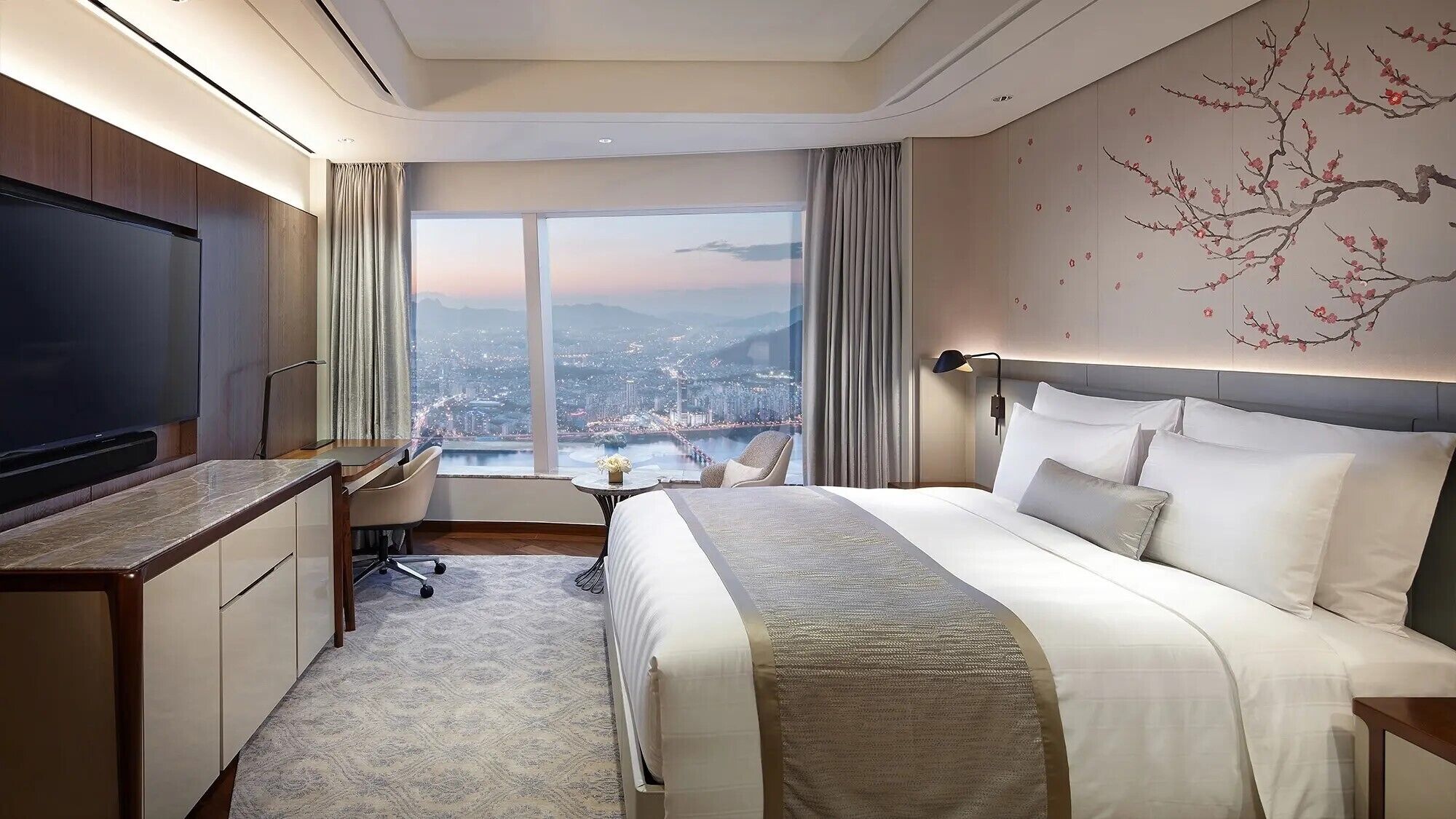 Una habitación de hotel moderna con una cama grande, un escritorio, una silla, un televisor y vistas a la ciudad a través de amplios ventanales.