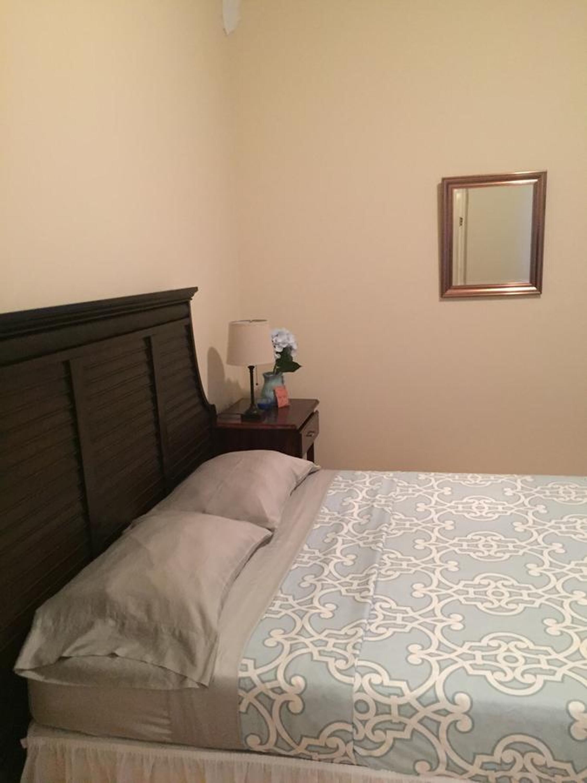A neatly made bed with a patterned bedspread, a wooden headboard, a bedside table with a lamp and a vase of flowers, and a mirror on the wall.