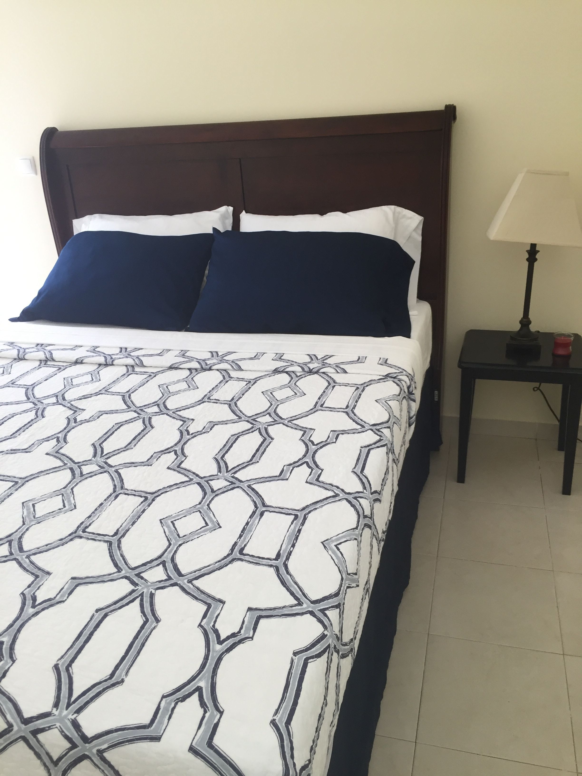 A bed with a geometric patterned bedspread, a dark wooden headboard, two blue pillows, and a small bedside table with a lamp and a red object.