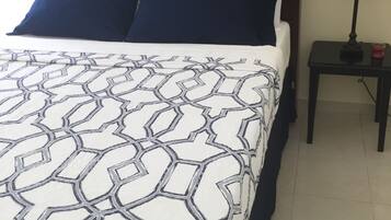 Una cama con una colcha de estampado geométrico, un cabecero de madera oscura, dos almohadas azules y una mesita de noche con una lámpara y un objeto rojo.