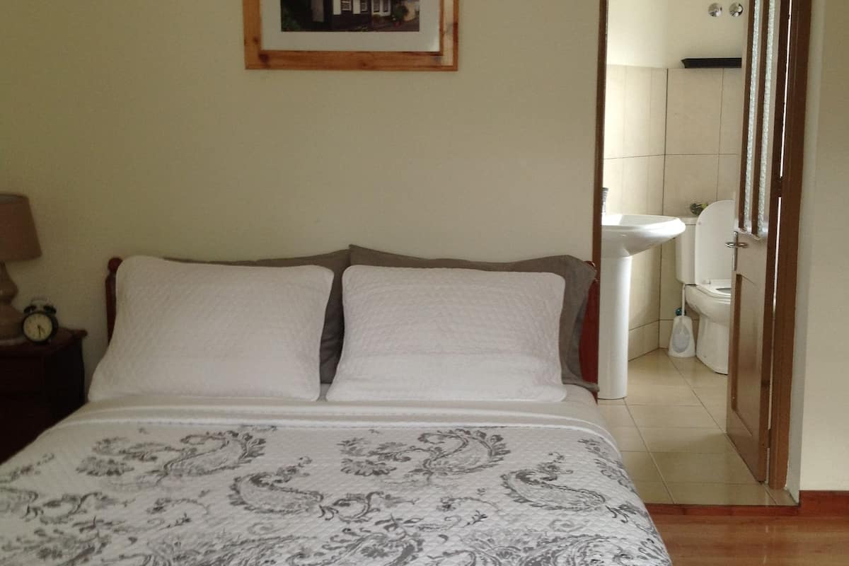 A bedroom with a bed, a nightstand with a clock, a framed picture on the wall, and a bathroom visible through an open door.