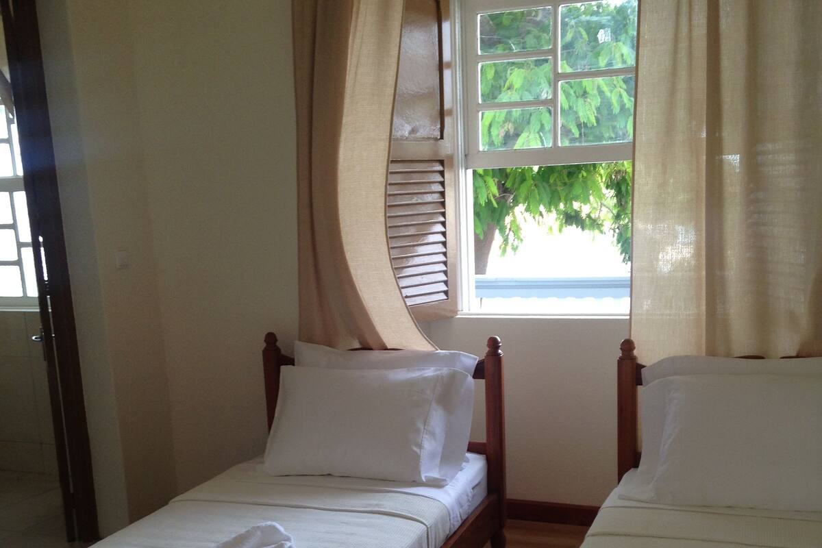 A room with two single beds, a window with blinds, and a view of greenery outside.
